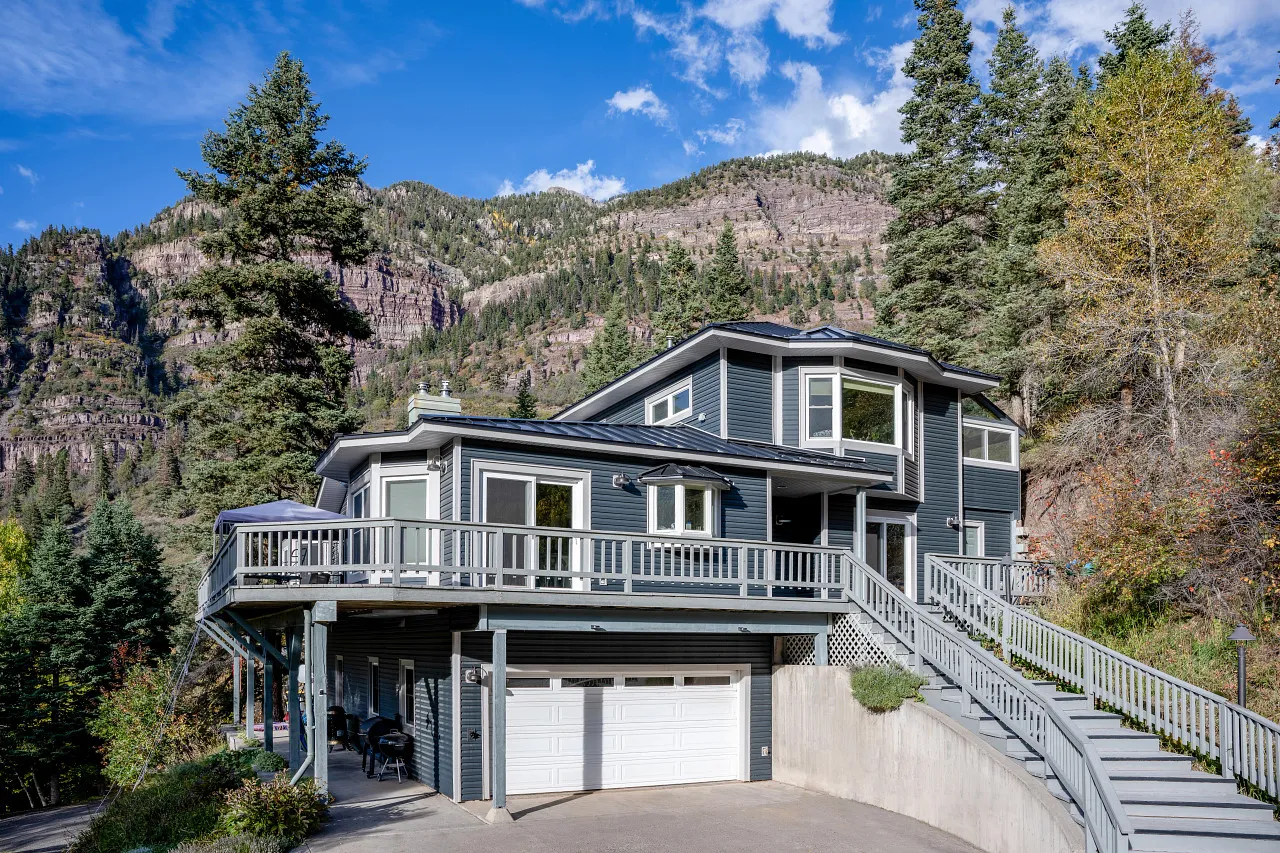 Modern two-story gray house with white trim, wraparound deck, and garage set against forested mountain backdrop under blue sky.